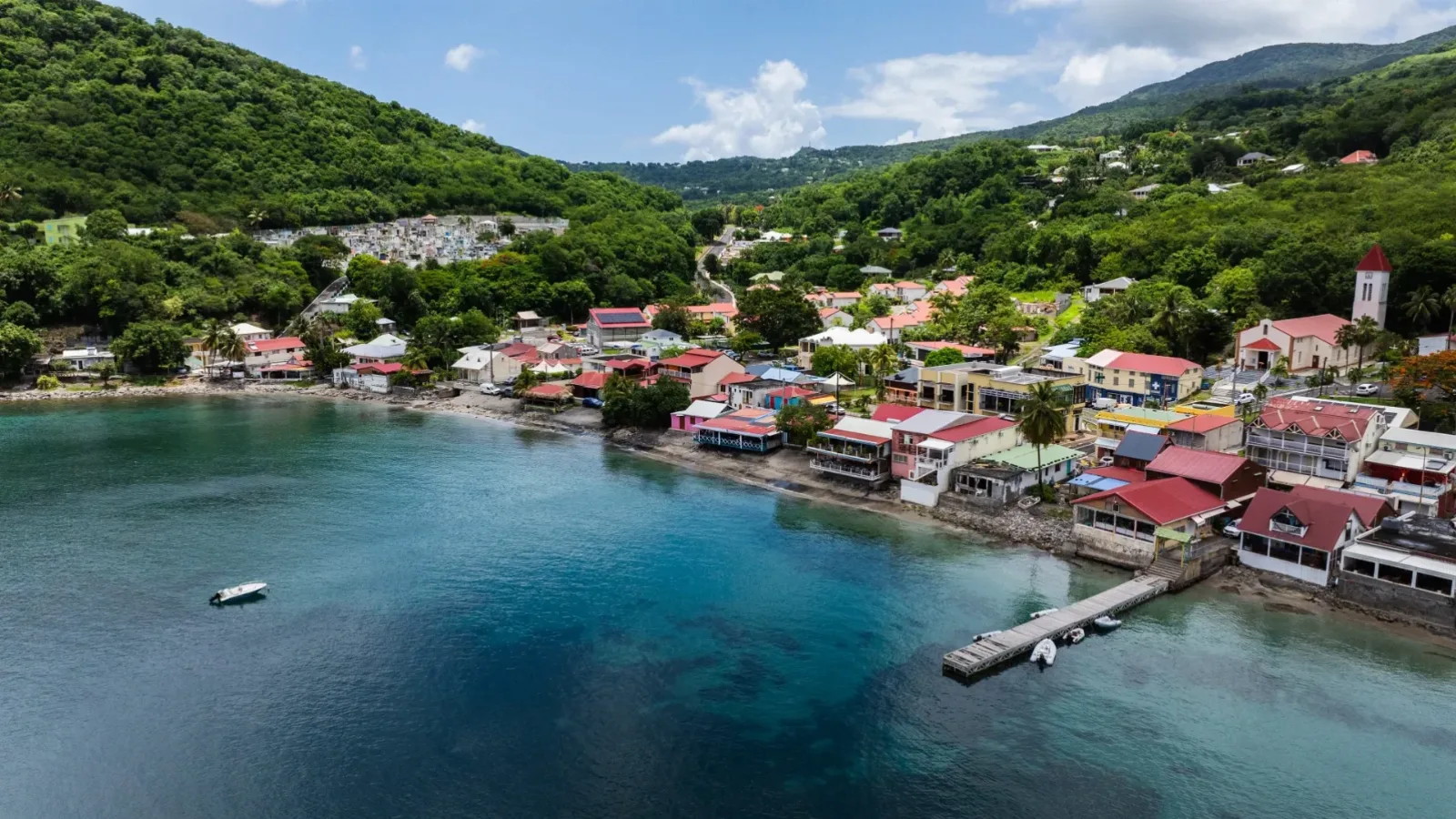 Vue aérienne du bourg de Deshaies au fond de sa baie, ses toits rouges, son ponton et les mornes boisés