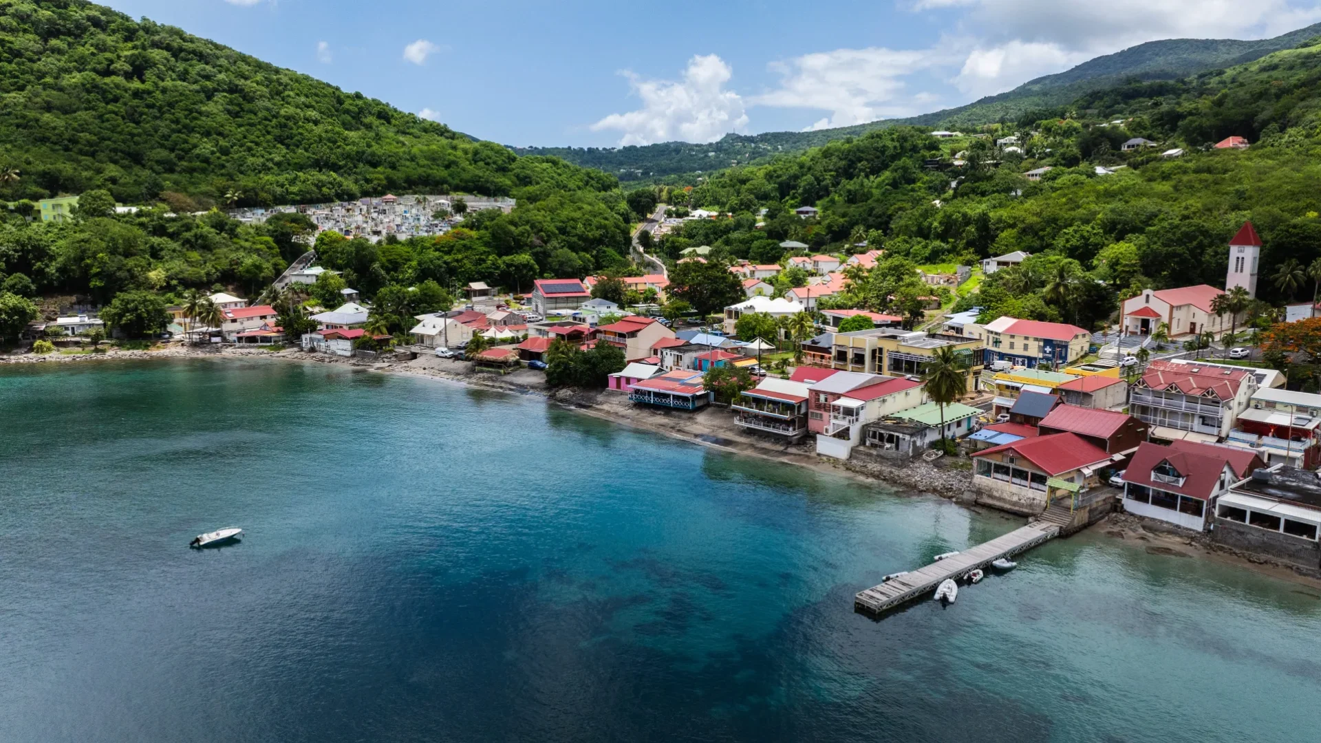 Vue aérienne du bourg de Deshaies au fond de sa baie, ses toits rouges, son ponton et les mornes boisés