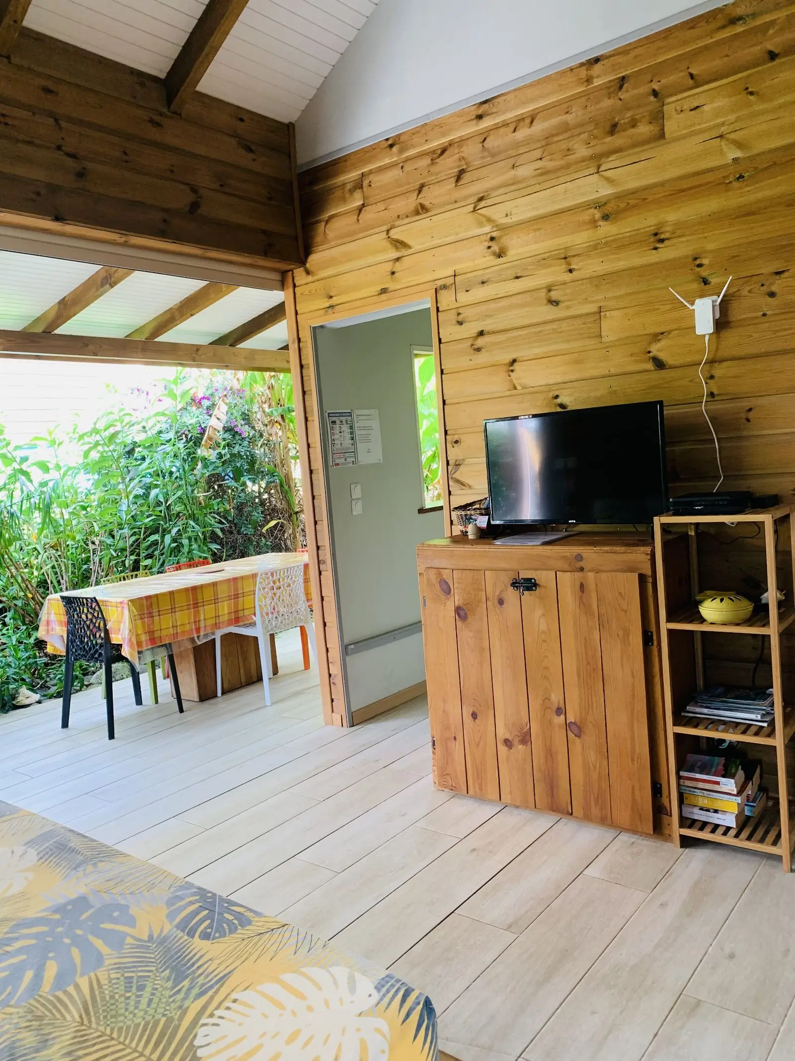 Séjour lambrissé de bois avec télévision et étagère, ouvert sur la terrasse et sa table