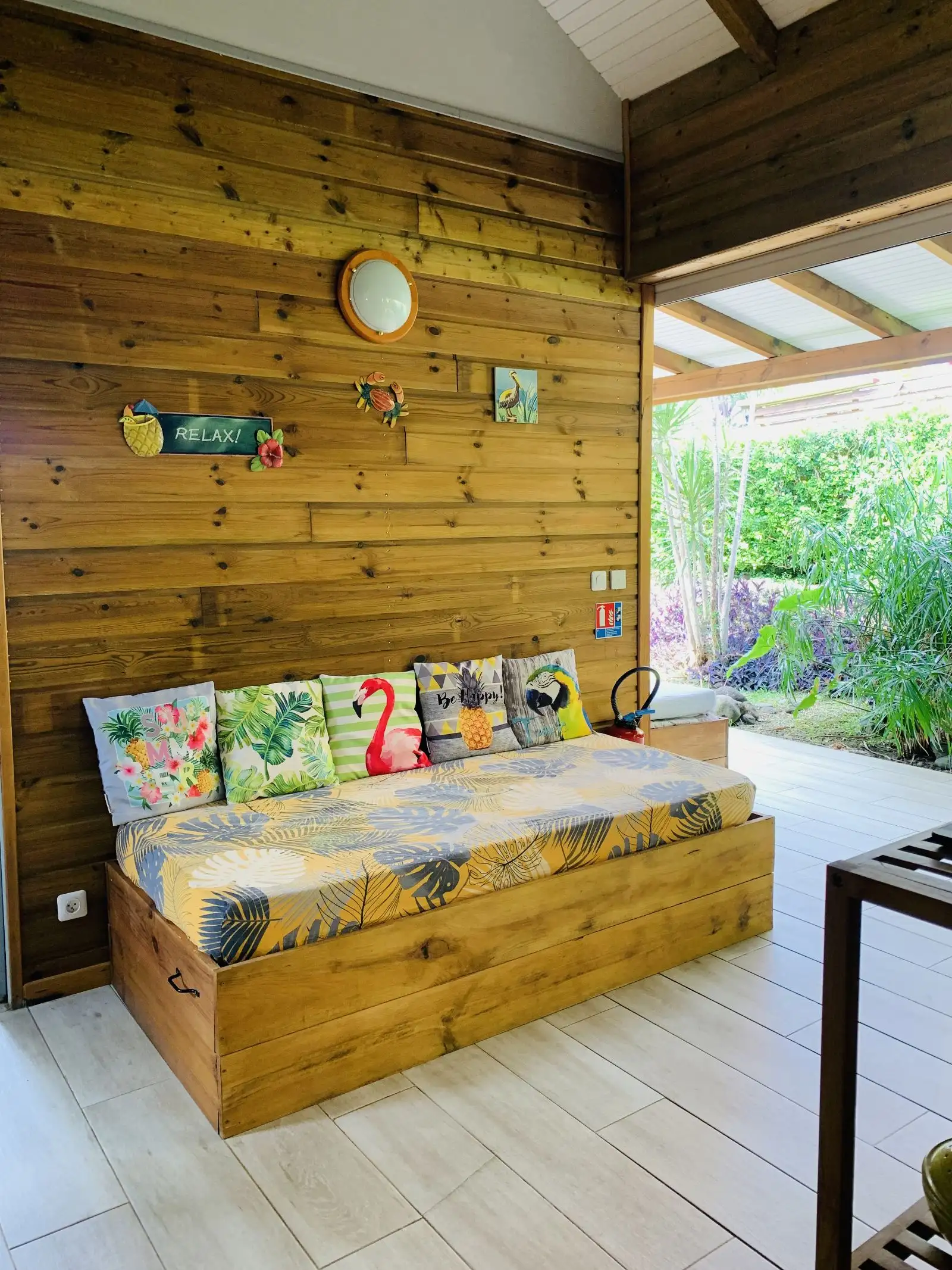 Banquette en bois garnie de coussins tropicaux, contre un mur lambrissé, face au jardin
