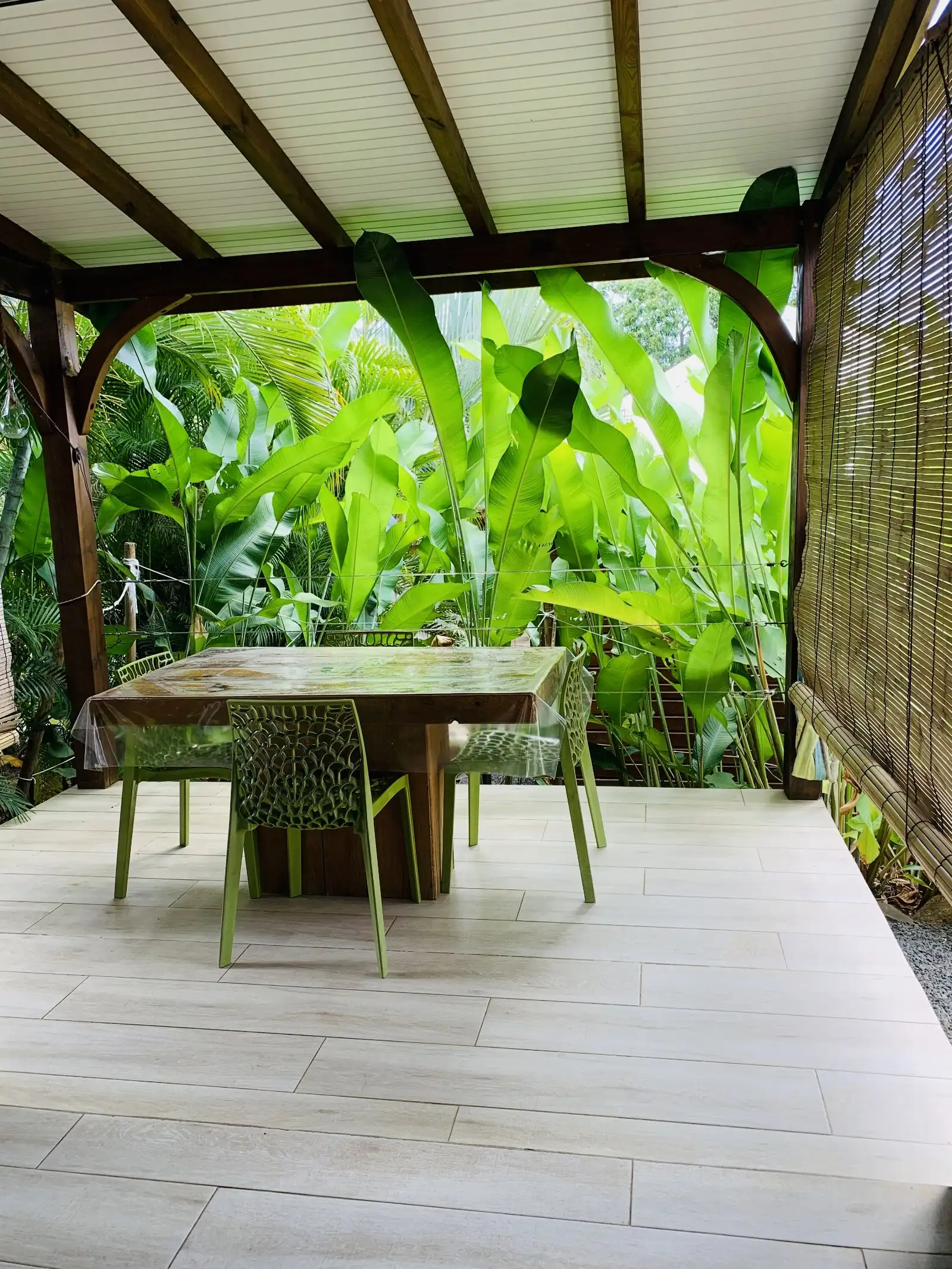 Terrasse couverte meublée d’une table en bois et de chaises vertes, face à des bananiers