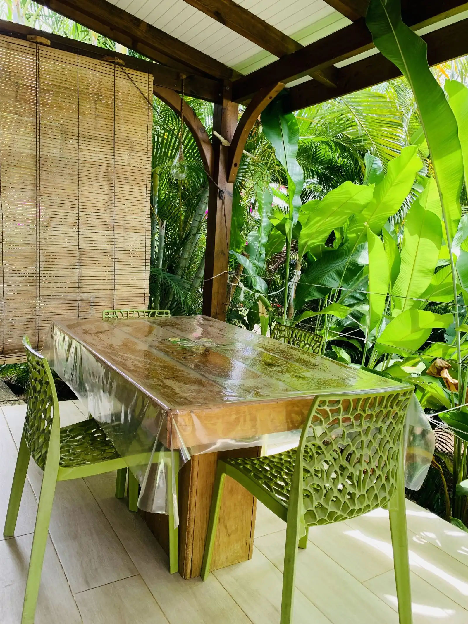 Table de repas en bois sur la terrasse, abritée par un store de bambou, ouverte sur la végétation
