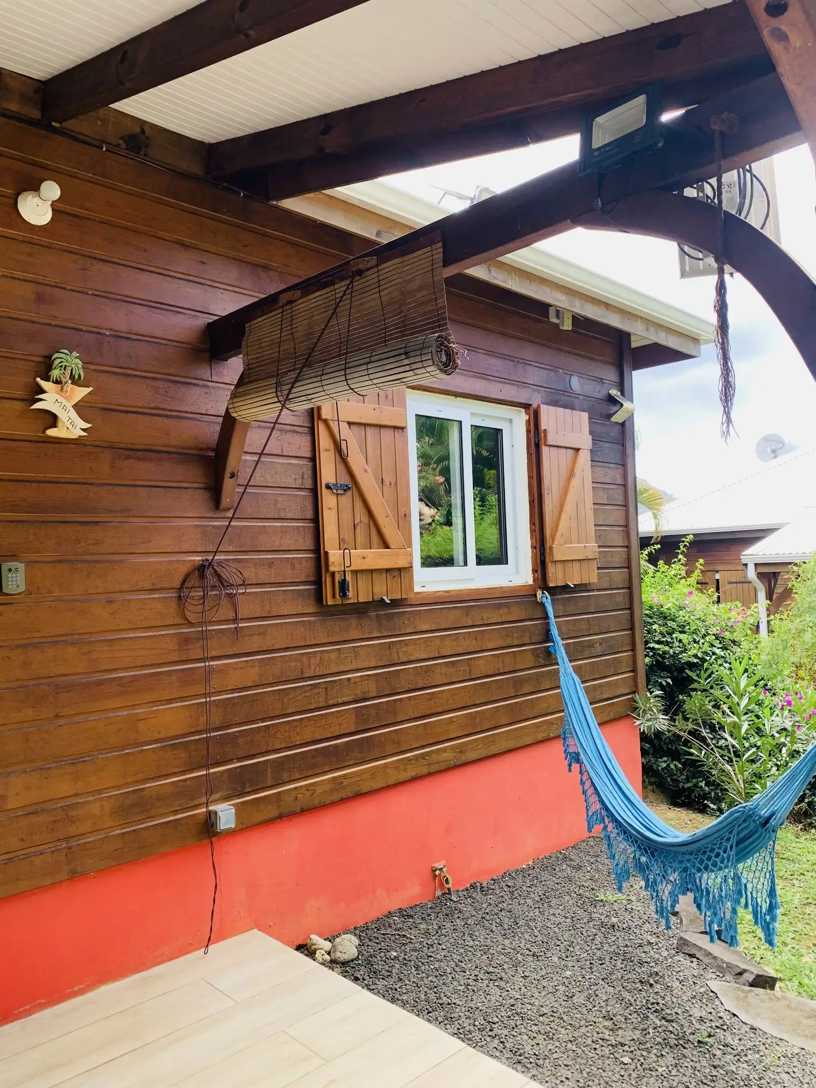 Façade en bois avec volets et hamac bleu, au-dessus d’un soubassement rouge