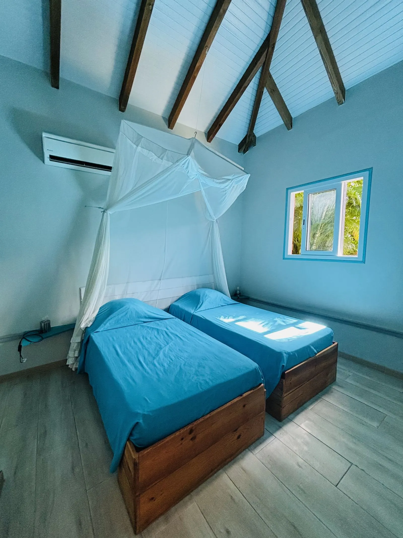 Chambre bleue avec deux lits simples sous moustiquaire, climatiseur et fenêtre sur la végétation